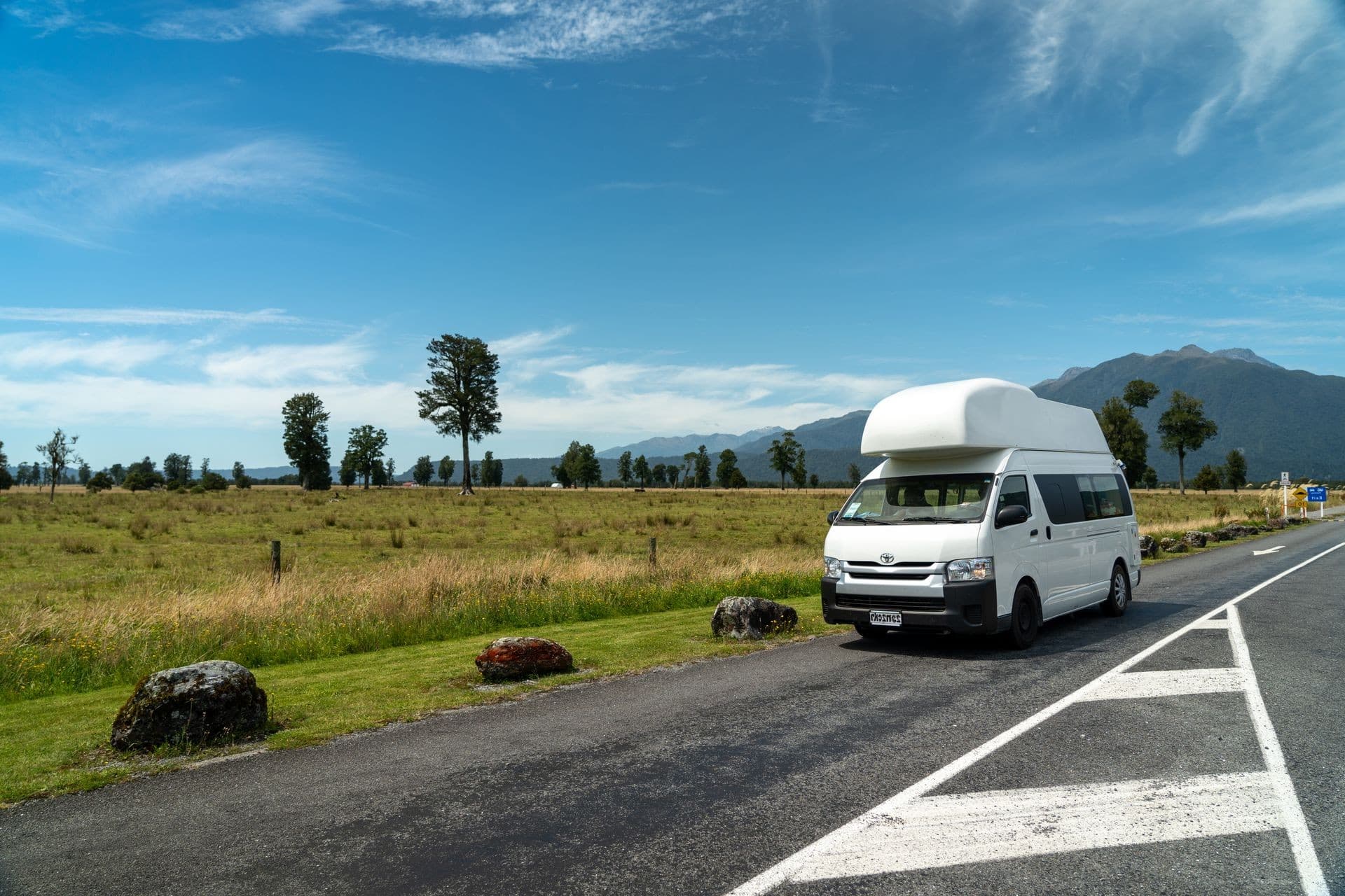 A camper van parked roadside beside an open field, mountains in the distance